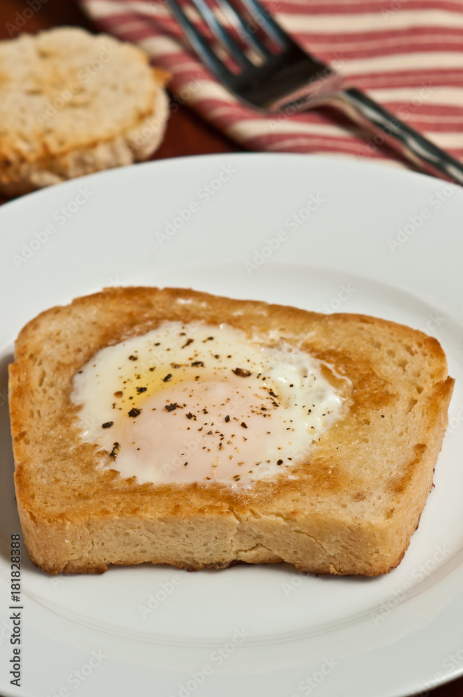 Single egg fried over light on white toast with the center  cut out, with fresh ground pepper, round, white dish, artisan fork and a red and white stripped towel in a basket 