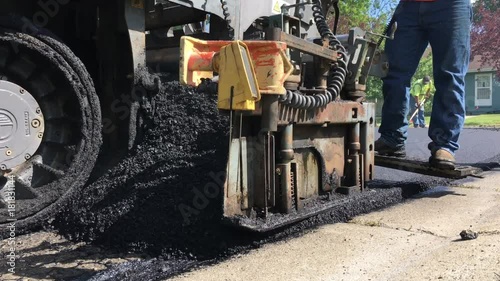 Asphalt laying machine with road construction worker on back putting down hot tar blacktop onto neighborhood street; shot from near ground level in bright sunlight.  