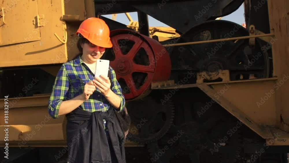 A young woman worker in orange helmet stands near a yellow old excavator and use a white telephone.