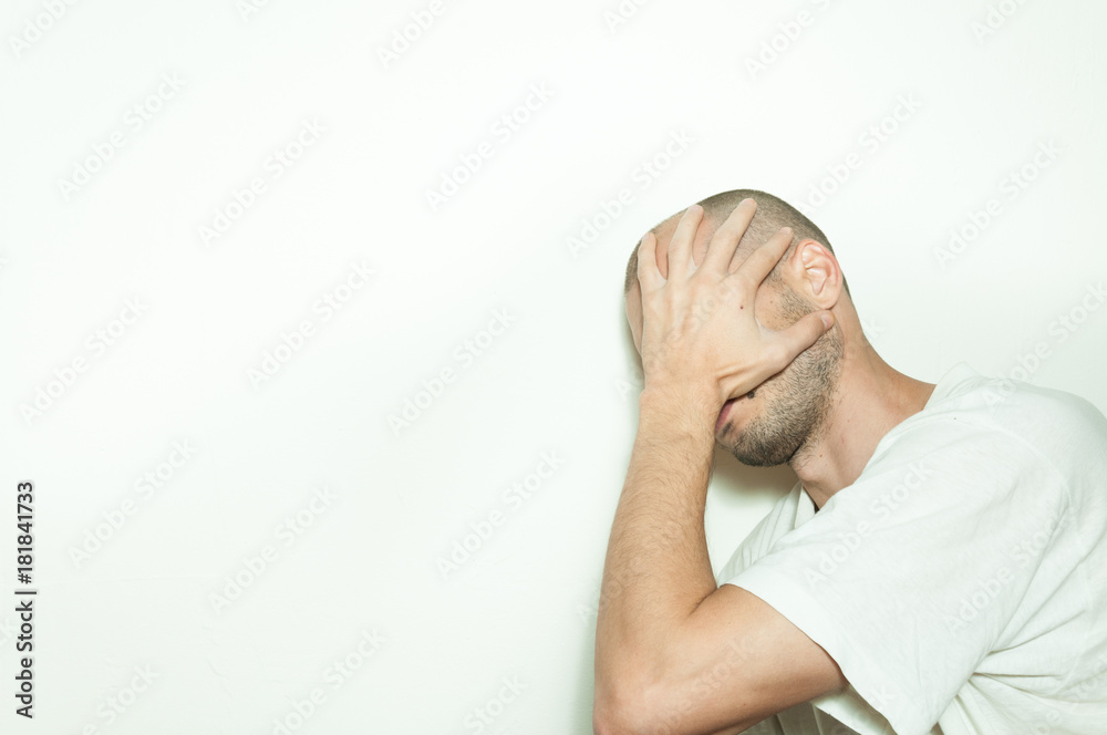 Young depressed man suffering from anxiety and feeling miserable cover his face with his hands and cry leaning on the white wall Young depressed man suffering from anxiety and feeling miserable cover his face with his hands and cry leaning on the white wall