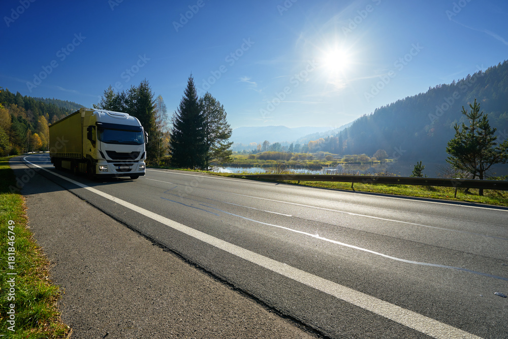 Glowing sun over the valley. White truck driving on the road around a lake and forested mountains in autumn.
