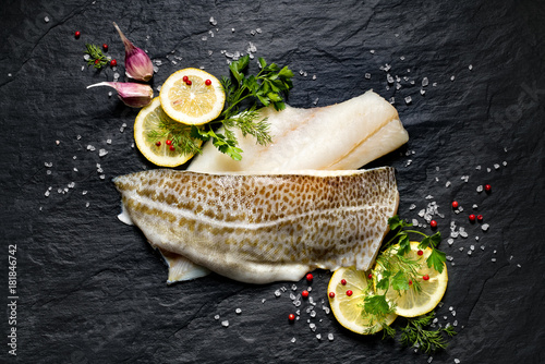 Fresh fish,  raw cod fillets with addition of herbs and lemon slices on black stone background, top view   photo