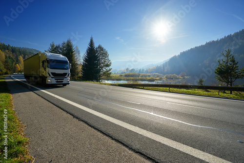 Obraz na płótnie Glowing sun over the valley. White truck driving on the road around a lake and forested mountains in autumn. w salonie