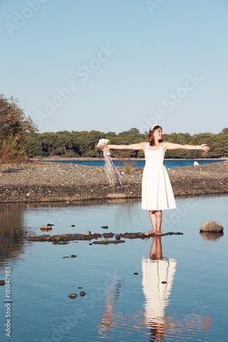 Happy young bride standing barefoot in lake shore with wedding dress and holding wedding bouquet and spread her arms and feeling freedom