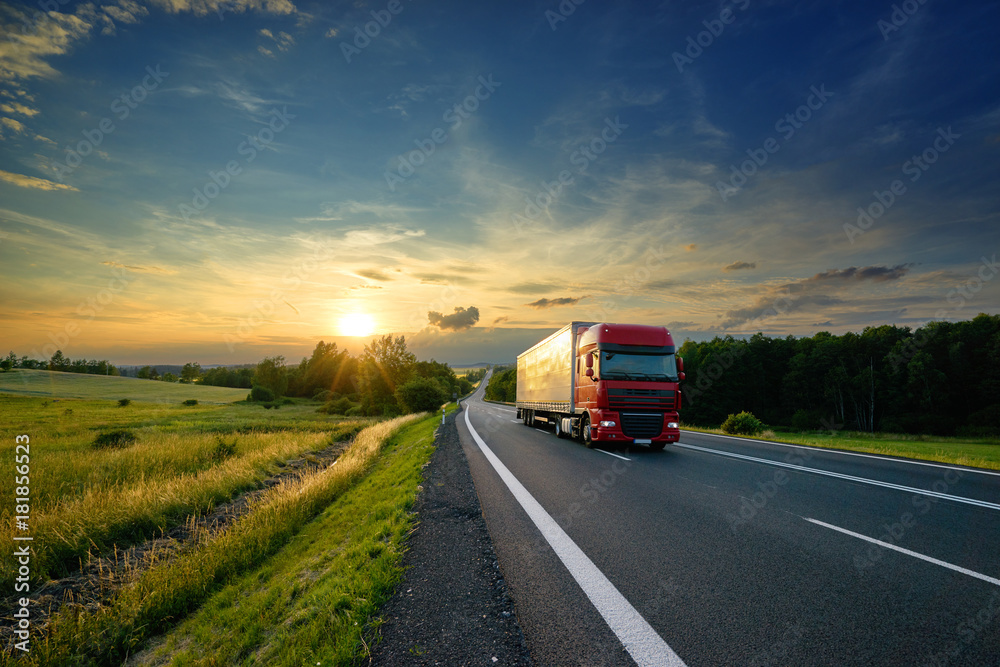 Red truck driving on the asphalt road in rural landscape at sunset