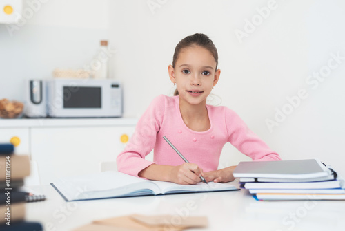 Girl doing homework in the kitchen. The dark-haired girl is dressed in a pink sweater.