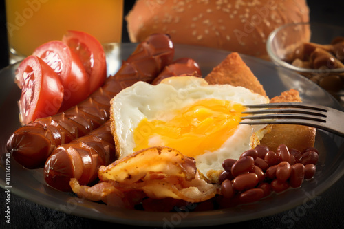 Full English breakfast with scrambled eggs, bacon, sausage, beans, tomatoes and juice. fork pierces the egg yolk. Black background