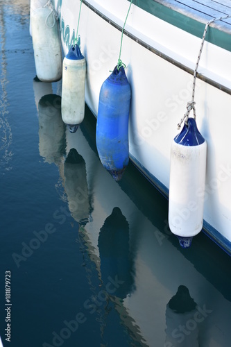 White and blue plastic fenders hanging on ropes along white ship hull.
