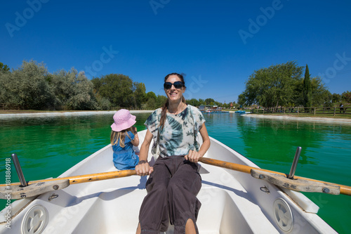 woman mother paddling in boat, smiling happy, next to four years old blonde girl, at park lake 