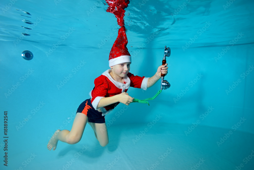 Little boy in Santa Claus hat floats under water in the pool and holding the phone in his hand. Portrait. Shooting under water. Horizontal orientation Little boy in Santa Claus hat floats under water in the pool and holding the phone in his hand. Portrait. Shooting under water. Horizontal orientation