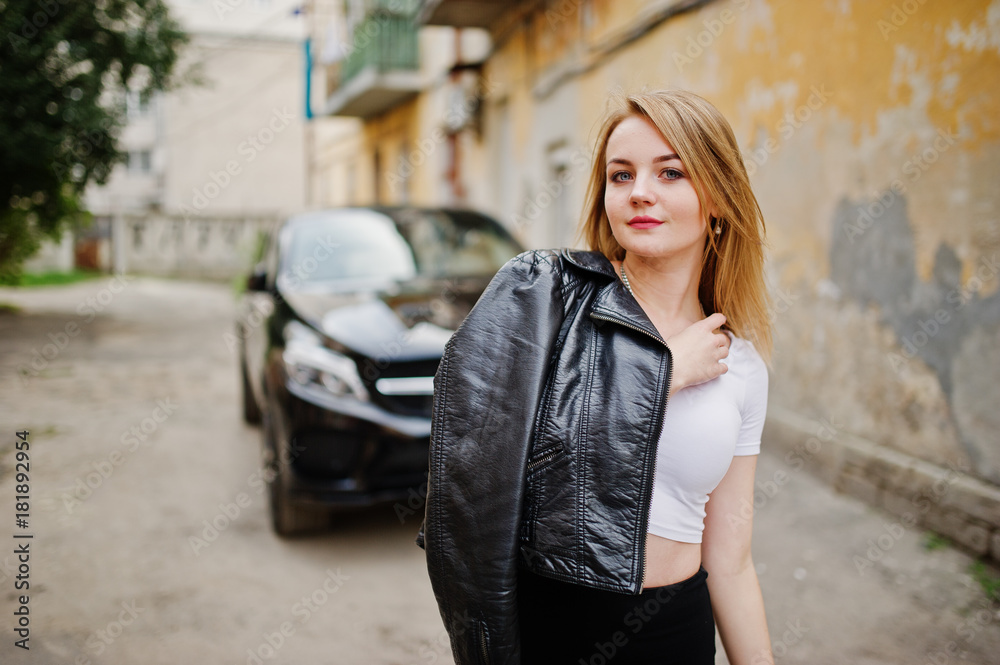 Elegant blonde girl wear on black leather jacket posing at streets of town background luxury car.