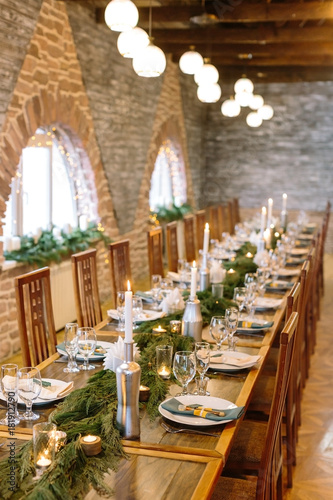 winter, meeting, holidays concept. extremely long wooden table in the restaurant hall for calebrating with lovely windows, there are lots of candles and aromatic conifer branches © melnikofd