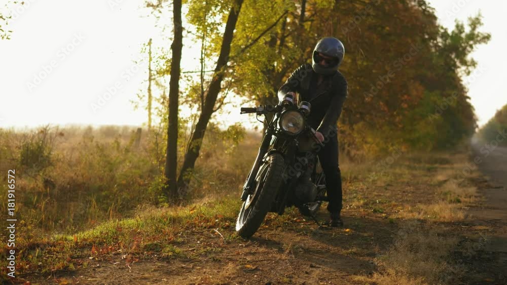 Back view of a man in helmet and leather jacket coming up to his bike and starting the engine while standing on the roadside in a sunny day in autumn Back view of a man in helmet and leather jacket coming up to his bike and starting the engine while standing on the roadside in a sunny day in autumn