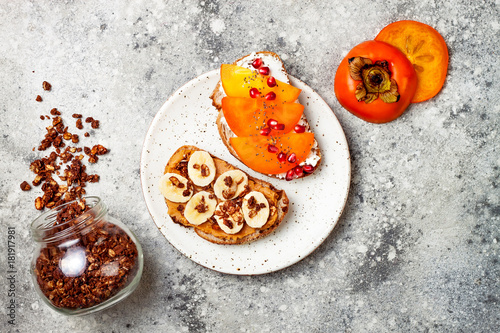 Healthy breakfast toasts with peanut butter, banana, chocolate granola, cream cheese, persimmon, pomegranate, chia seeds. Top view, overhead, copy space