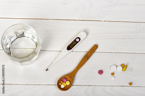 table spoon with tablets, thermometer and glass of water on white wooden table. Medical concept photo