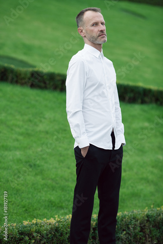 Luxurious and stylish at any age. Portrait of blue eyed rich mature man posing over green lawn background. Premium class white shirt. Classic style. Outdoor shot