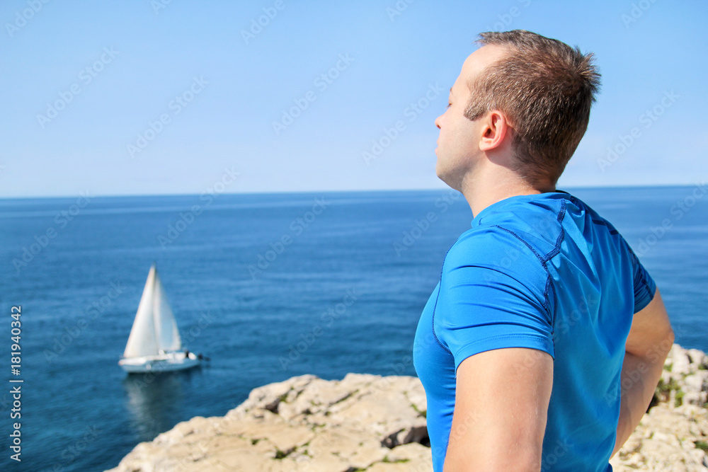 Handsome man wearing a sports clothing takes a break after a running training and enjoys beautiful natural surroundings, overlooking the horizon of the Adriatic sea, a sailboat sailing in background.