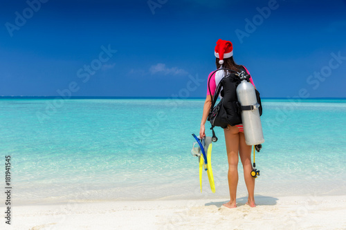 Taucherin mit Ausrüstung und Weihnachtsmütze steht am tropischen Strand und schaut auf den Ozean