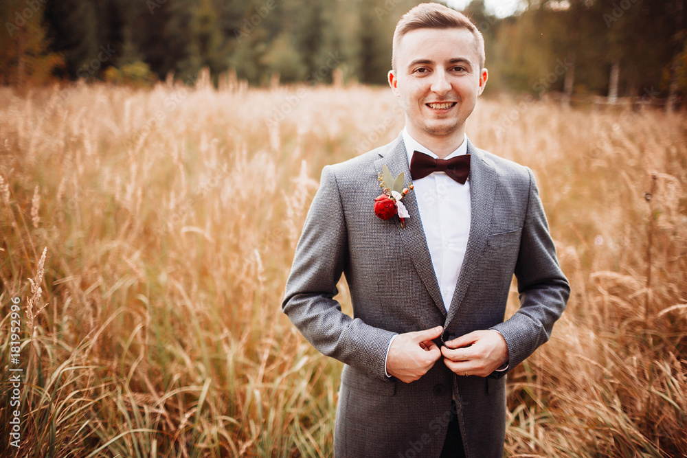 Handsome groom in grey jacket stands on golden field Handsome groom in grey jacket stands on golden field