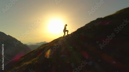 Wallpaper Mural AERIAL SILHOUETTE: Flying over young woman hiker as she climbs up sloping terrain on her way to summit. Woman trying to reach mountain peak before sundown. Lady climbing uphill in beautiful scenery. Torontodigital.ca