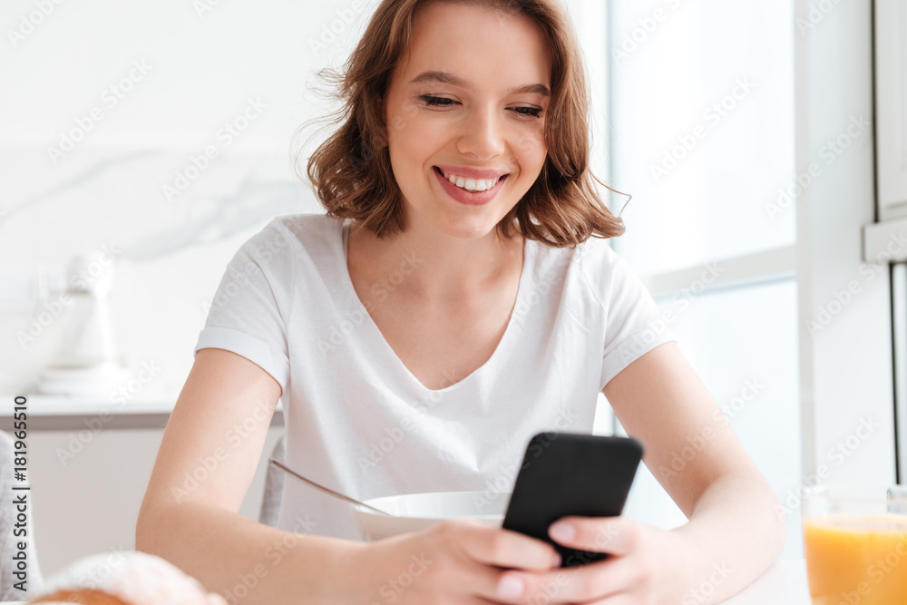 Close-up portrait of joyful pretty girl texting message on smartphone while sitting and having breakfast at the kitchen Close-up portrait of joyful pretty girl texting message on smartphone while sitting and having breakfast at the kitchen