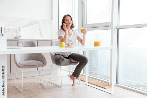 Young attractive woman in casual wear speaking on mobile phone and holding glass of orange juice while sitting at kitchen table