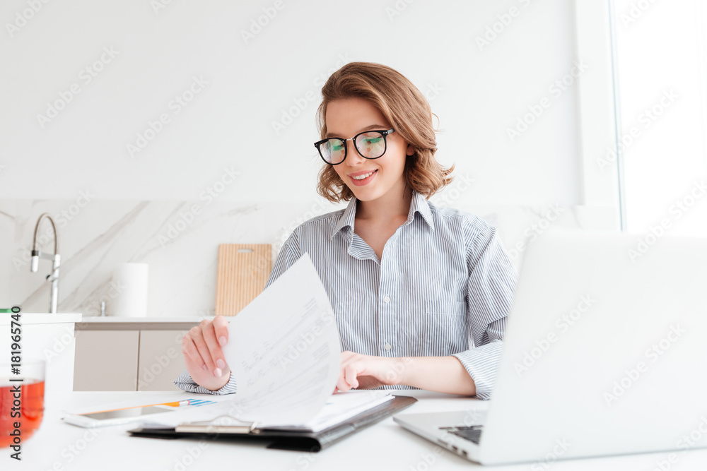 Photo of cheerful attractive woman in glasses reading new contract while working in the kitchen Photo of cheerful attractive woman in glasses reading new contract while working in the kitchen