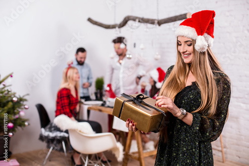 Portrait of a young woman with a gold giftbox at home on the foreground. Beautiful blonde in a Santa hat and black dress