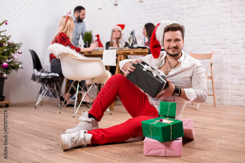 Shot of happy friends enjoying holidays. Focus on the man with a gift boxes in the foreground in a red Christmas hat