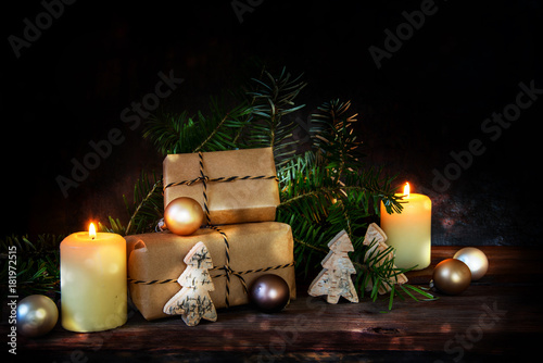 gift boxes in kraft paper and two burning candles decorated with christmas balls, fir branches and small wooden trees on a dark rustic background with copy space