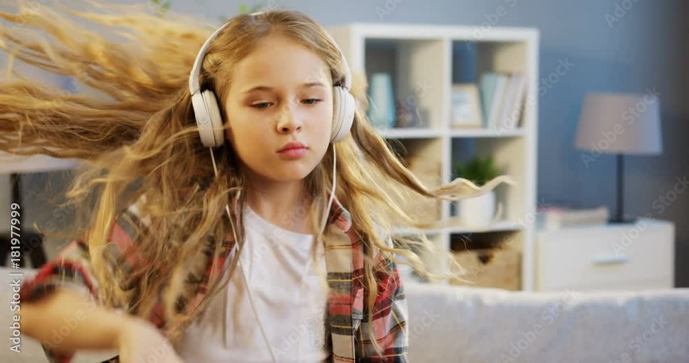Portrait shot of lovely blonde girl in the big white headphones listening to the music and moving to the rhythm in the nice room at home. Inside Portrait shot of lovely blonde girl in the big white headphones listening to the music and moving to the rhythm in the nice room at home. Inside