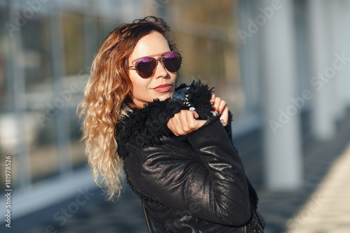 woman in sun glasses a black leather jacket, black jeans posing in front of mirrored windows. Female fashion concept. Outdoor.
