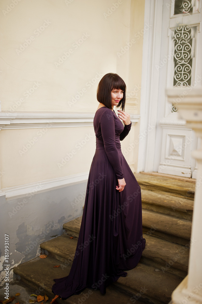 Adult brunette woman at violet gown background old vintage house with stairs. Adult brunette woman at violet gown background old vintage house with stairs.