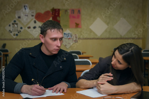 Two students sit at a desk and write an examination work. The male student looks on female student notebook and tries to write off the exam work but the student girl closes the notebook with her hand.