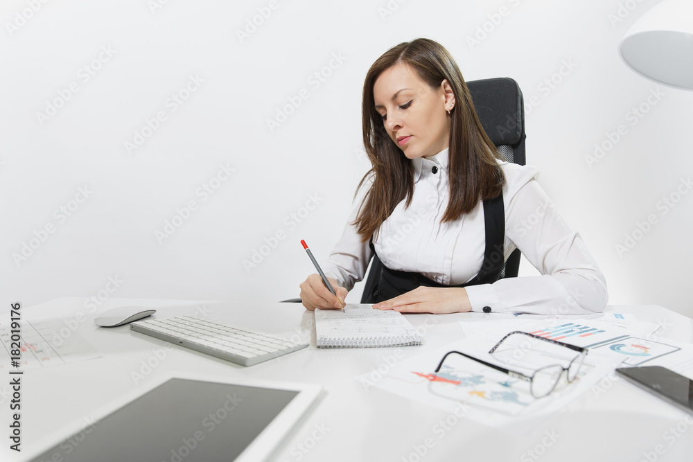 Beautiful serious brown-hair business woman in suit and glasses sitting at the desk, working at computer with modern monitor and documents in light office, writing in notebook on white background Beautiful serious brown-hair business woman in suit and glasses sitting at the desk, working at computer with modern monitor and documents in light office, writing in notebook on white background