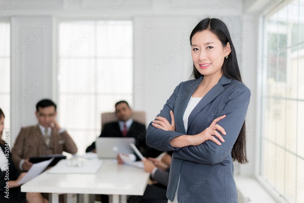 Happy businesswoman standing with arms crossed at office while colleagues discussing in background. Happy businesswoman standing with arms crossed at office while colleagues discussing in background.