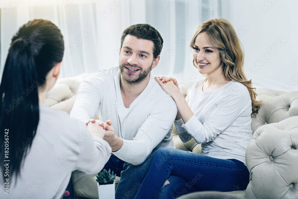 Fototapeta premium You saved our family. Joyful delighted handsome man holding a hand of his psychologist and expressing his gratitude while sitting with his wife on the sofa
