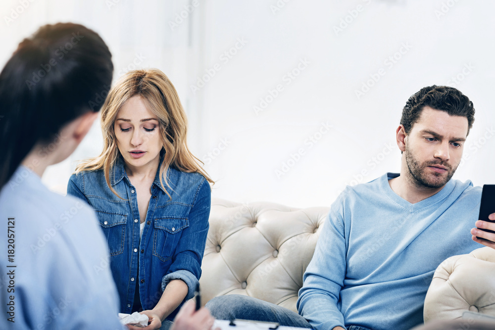 Obraz premium I dont care. Handsome bearded young man playing games on his smartphone and showing no interest in psychological session while sitting with his wife on the sofa