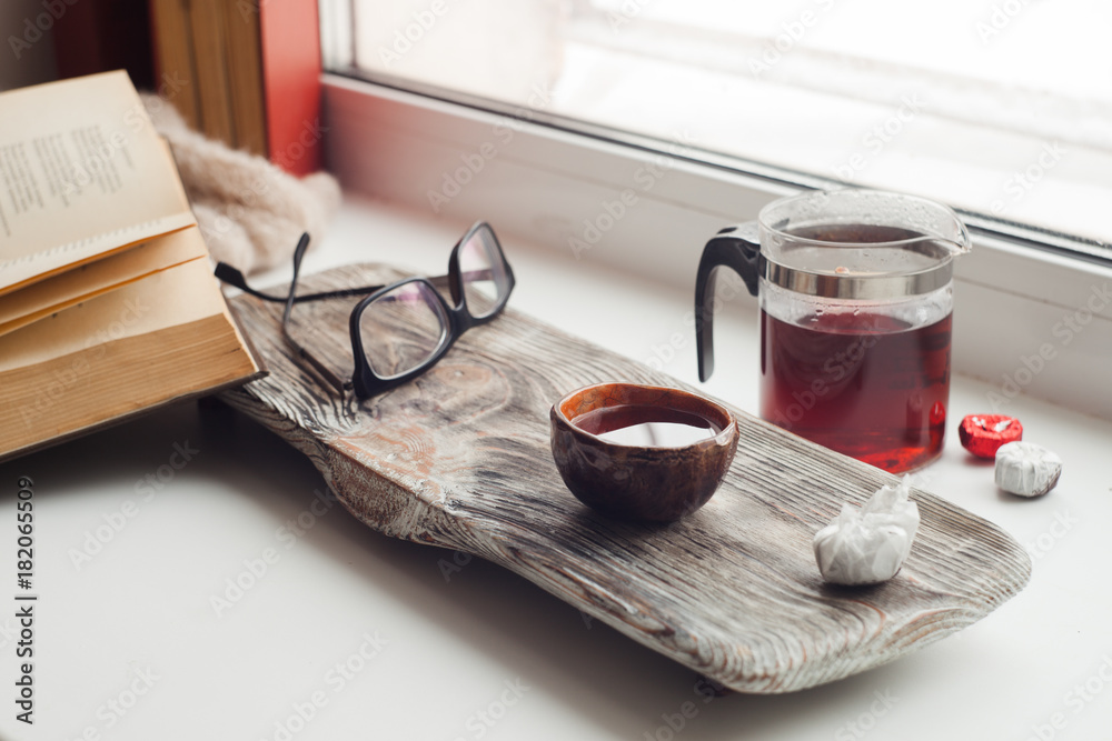 Still life details, book, glasses, cup of asian tea and teapot on vintage wooden tray on windowsill in living room. Lazy winter weekend with a book at home Still life details, book, glasses, cup of asian tea and teapot on vintage wooden tray on windowsill in living room. Lazy winter weekend with a book at home