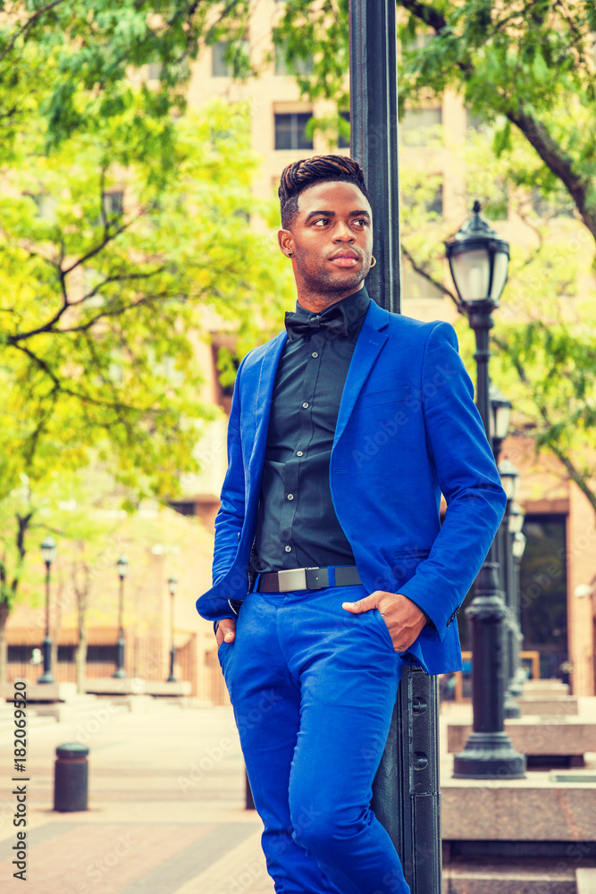 African American Man Fashion in New York. Dressing formally in blue suit,  black shirt, bow tie, wearing earring, black man with afro hairstyle stands  against light pole on street, looks forward. Stock