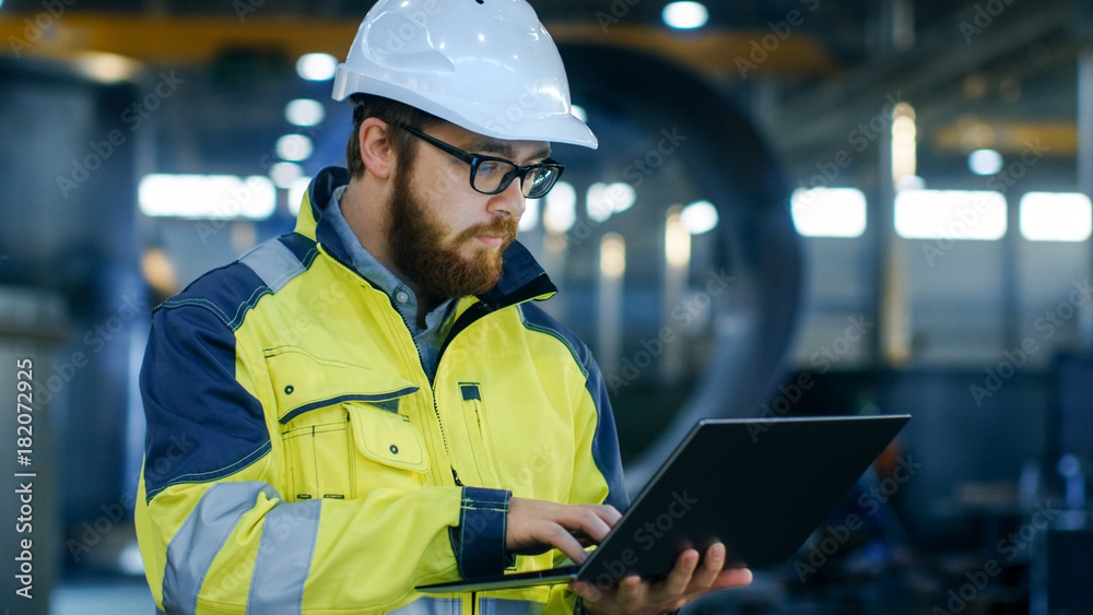 Obraz premium Industrial Engineer in Hard Hat Wearing Safety Jacket Uses Laptop. He Works in the Heavy Industry Manufacturing Factory with Various Metalworking Processes are in Progress.
