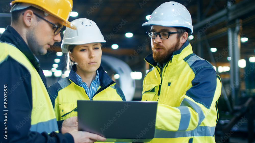 Male and Female Industrial Engineers Talk with Factory Worker while Using Laptop. They Work at the Heavy Industry Manufacturing Facility. Male and Female Industrial Engineers Talk with Factory Worker while Using Laptop. They Work at the Heavy Industry Manufacturing Facility.