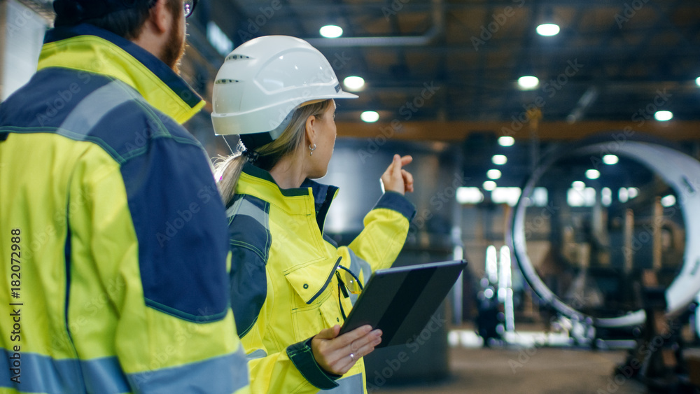 Male and Female Industrial Engineers in Hard Hats and Safety Jackets Discuss New Project while Using Tablet Computer. They Work at the Heavy Industry Manufacturing Factory. Male and Female Industrial Engineers in Hard Hats and Safety Jackets Discuss New Project while Using Tablet Computer. They Work at the Heavy Industry Manufacturing Factory.