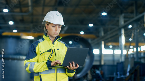 Fotografie Female Industrial Engineer in the Hard Hat Uses Laptop Computer while Standing in the Heavy Industry Manufacturing Factory