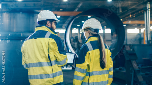 Male and Female Industrial Engineers use Laptop and Have Discussion While Walking Through Heavy Industry Manufacturing Factory. They Wear Hard Hats and Safety Jackets.