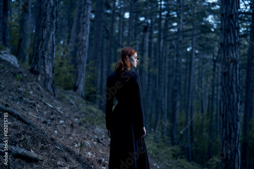 Young beautiful woman in a long black dress is standing in a dark forest