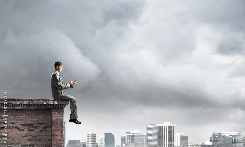 Man on roof edge send message with smartphone and cityscape at background Man on roof edge send message with smartphone and cityscape at background