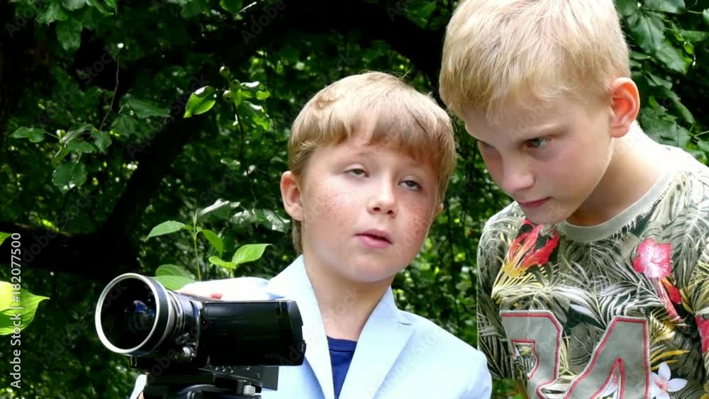 Young boys with video camera shoots film about nature of green park background. Children outdoors in summer are creative work of cinema. Beautiful footage. Interesting to look at world in childhood.