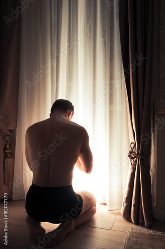 Young adult praying man sits near window