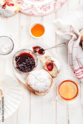 Tea time with festive sufganiyot donuts filled with jelly and covered with sugar powder. White wooden background, bright lighting. Vertical composition.